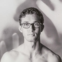 black and white portrait of young man with glasses, blurred hand pattern in background.