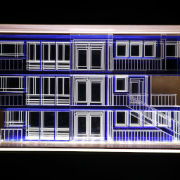 3D geometric acrylic model, rectangular windows and blue features, in a glass case with wooden back under LED lights.