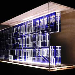 3D geometric acrylic model, rectangular windows and blue features, in a glass case with wooden back under LED lights.