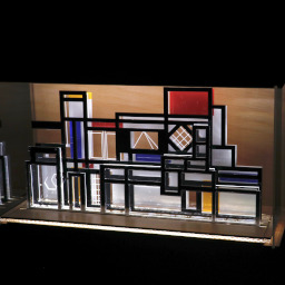 3D geometric acrylic model, various rectangular features and red, blue and yellow features, in a glass case with wooden back under LED lights.
