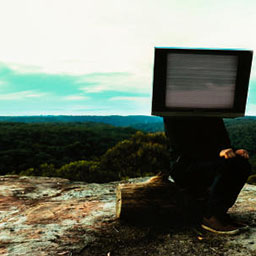 figure of man sitting on wooden log, TV on head, in rocky landscape.