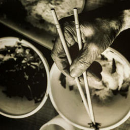close-up wrinkled hand holding chopsticks, reaching down to two bowls of food, black and white photograph.
