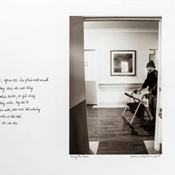 old woman walking along hallway with walking frame, dark floorboards, cursive writing on the left, black and white photograph.
