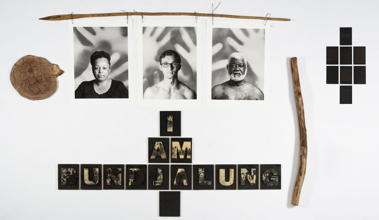 a collection of wooden objects,a tree stump, spear and didgeridoo, surrounded by three portraits and large single letters spelling out I AM BUNDJALUNG.