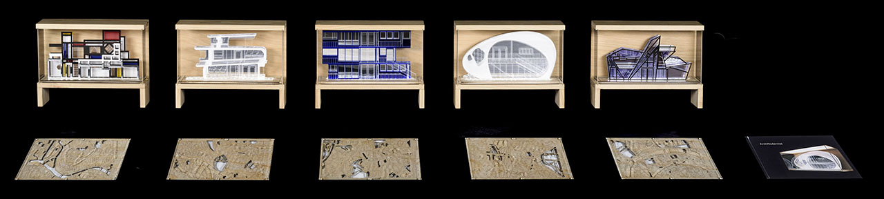 five wooden boxes with models of modern geometric architecture, five flat wooden panels of designs below and one black book lying flat to the right.