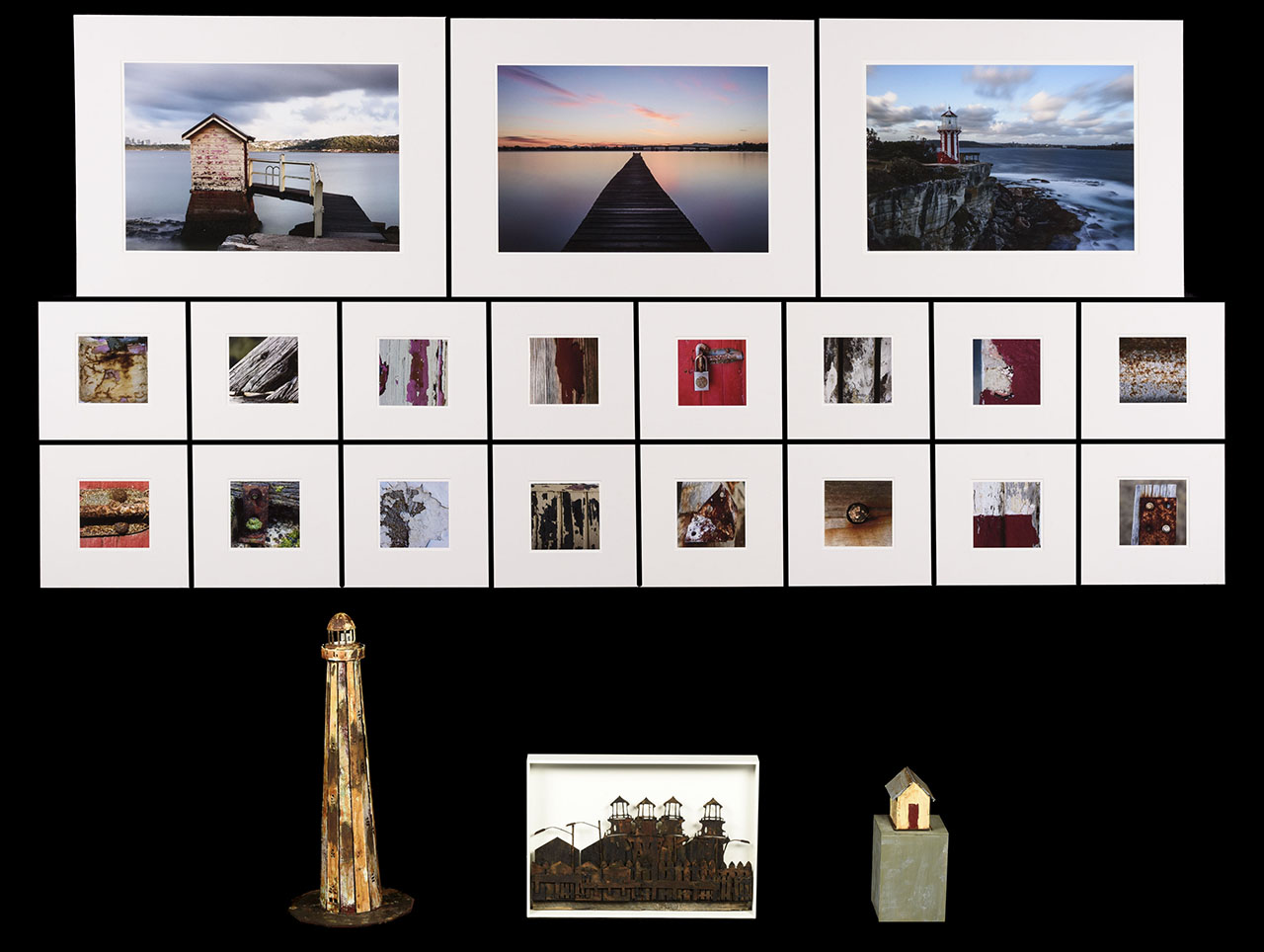 three large photographic prints of oceanic landscapes with man-made elements, sixteen small close-up photographs of wood and metal detailing and three individual sculptures of a long lighthouse, rusty collection of houses and isolated house on a wooden block.