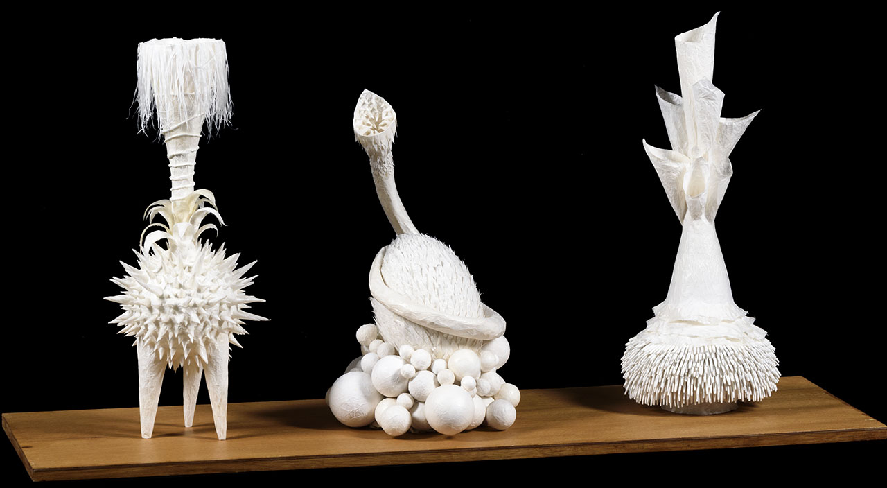 three tall white ceramic sculptures with curved lines and textural detail sitting on wooden shelf.