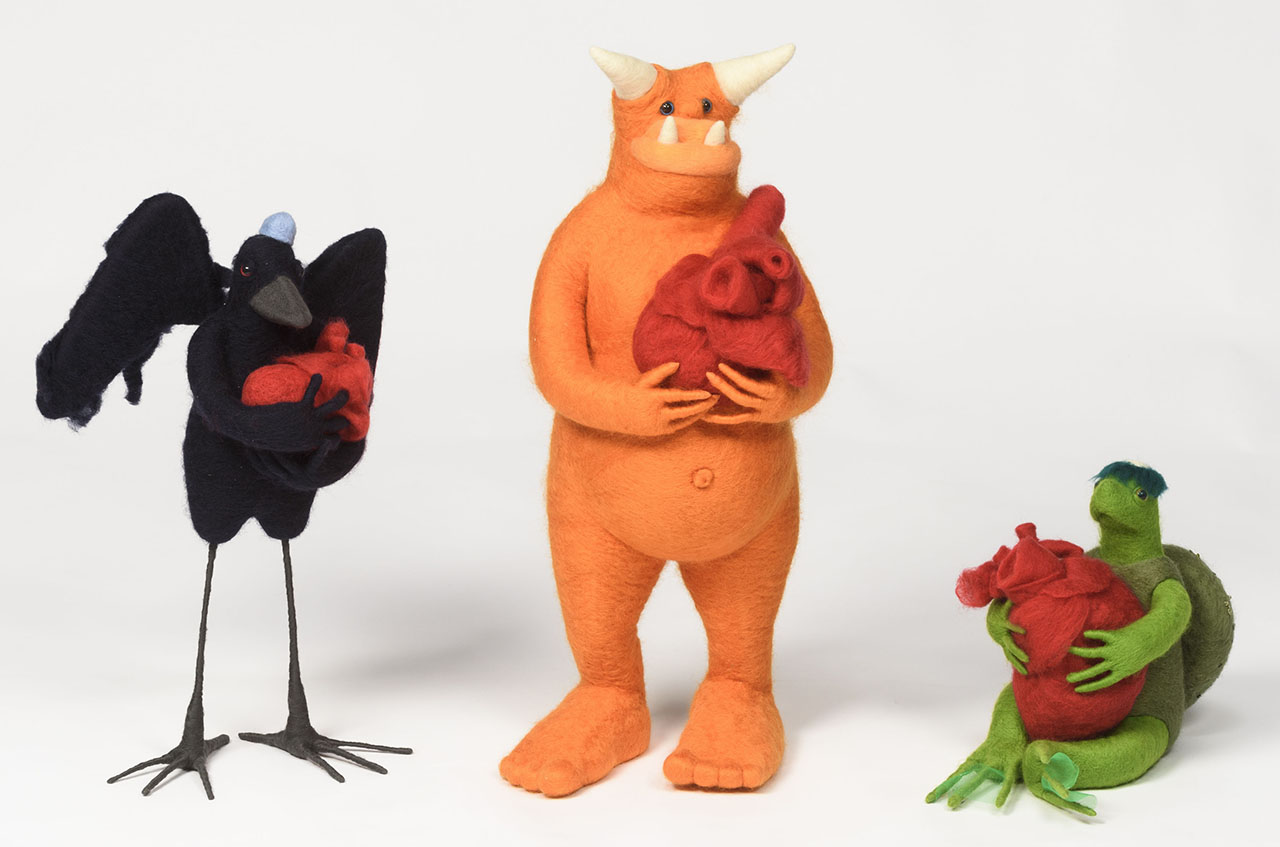 three felt sculptures, one black raven, one orange monster and one green turtle, all holding red hearts.
